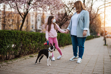 Mother and daughter with protective masks on their faces enjoying in walk in park with their dog.