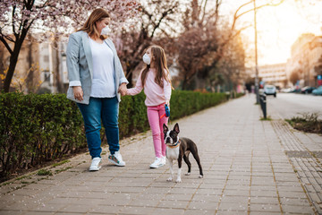 Mother and daughter with protective masks on their faces enjoying in walk in park with their dog.