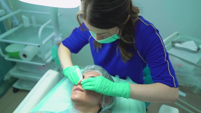 skilled cosmetologist in uniform squishes pimples on young woman face in modern cosmetic salon view from above