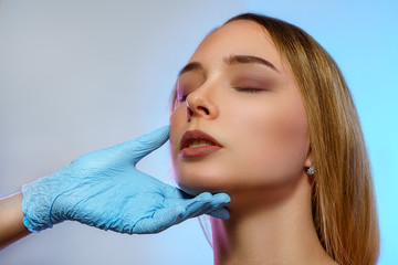 Doctor's hands in gloves touch the face of young woman isolated on blue background. Healthcare, cosmetology, medical and plastic surgery concept