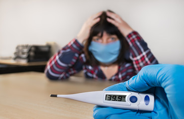 Pandemic Covid-19. High fever on electronic medical thermometer in nurse hand in glove. Sick woman with blue face mask in doctor office. Headache and sore throat. Fear of coronavirus disease outbreak.