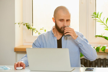 A man drinking a coffee while working remotely on his laptop during the quarantine to avoid the...