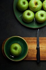 Green apples on a green plate.  A single apple is isolated on a smaller plate and placed on a wooden board next to a knife.  Flatlay against a dark grey background.