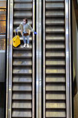 Obraz premium Woman with yellow luggage stands on escalator at almost empty airport terminal due to coronavirus pandemic/Covid-19 outbreak travel restrictions. Flight cancellation. Quarantine measures. Top view. 