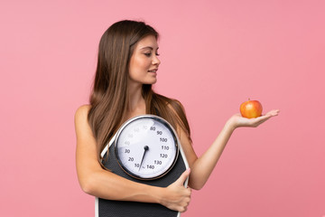 Young girl with weighing machine and with an apple over isolated pink background