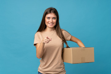 Young woman over isolated blue background holding a box to move it to another site while pointing to the front
