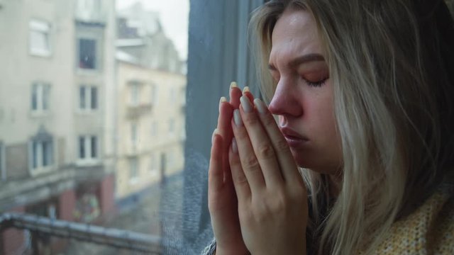 Upset young miserable woman suffers fever or cold looks through window coughing and sneezing. Unhappy girl staying at home drawing a sad face on window waiting isolation period. Coronavirus.