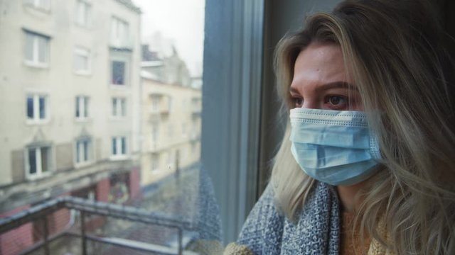 Sick cold young woman wearing protective white mask coughing badly looking in window quarantied due to coronavirus outbreak. Health care. COVID-19 isolation period.