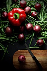Green beans and red onions with red pepper scattered across a dark grey background. A wooden chopping board with a knife with a black handle sits next to a onion. Portrait flat lay or top view.