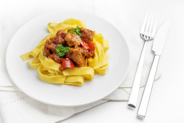 Stew or goulash or from pork meat with onions and red peppers served on tagliatelle noodles, plate,...