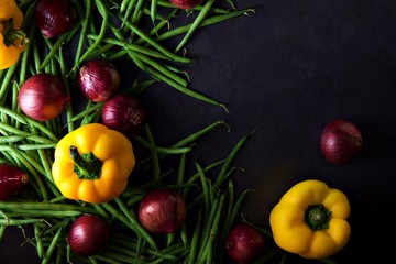 Green beans and red onions with yellow peppers scattered across a dark grey background. 
