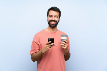 Young man with beard holding a take away coffee over isolated blue background sending a message with the mobile