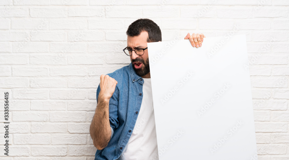 Wall mural Handsome man with beard over white brick wall holding an empty placard