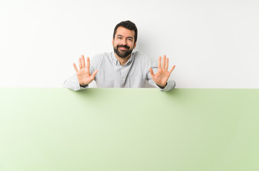 Young handsome man with beard holding a big green empty placard counting ten with fingers