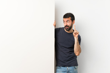 Young handsome man with beard holding a big empty placard intending to realizes the solution while lifting a finger up