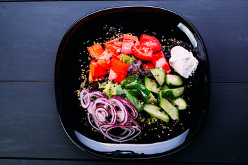 Healthy vegetable salad of fresh slices tomato, cucumber, onion rings, cabbage leaf and white sauce on a black plate. Diet menu. top view, blue wooden background