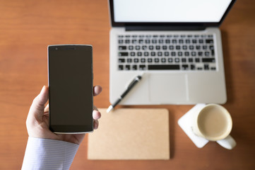 Young businessman holds in his hand a smart phone and in an unfocused background a wooden desk with a laptop, a notebook and a cup of coffee. Business and lifestyle concept