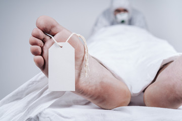 Feet of a dead body, with an identification tag - blank sign attached to a big toe. Covered with a white sheet. Coronavirus victim.