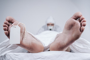 Feet of a dead body, with an identification tag - blank sign attached to a big toe. Covered with a white sheet. Coronavirus victim.