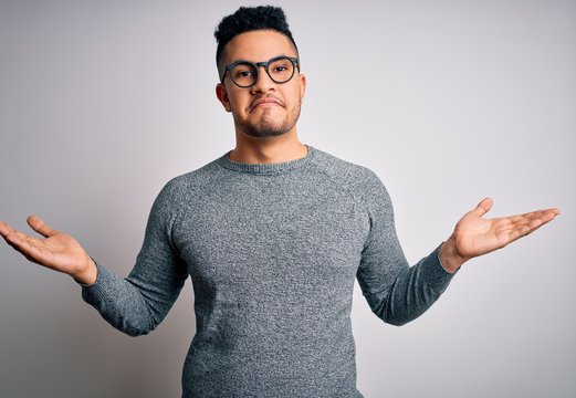 Young handsome man wearing casual sweater and glasses over isolated white background clueless and confused expression with arms and hands raised. Doubt concept.
