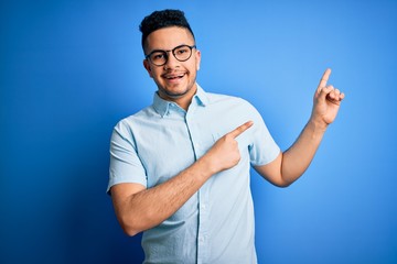 Young handsome man wearing casual summer shirt and glasses over isolated blue background smiling and looking at the camera pointing with two hands and fingers to the side.