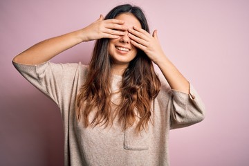 Young beautiful brunette woman wearing casual sweater standing over pink background covering eyes with hands smiling cheerful and funny. Blind concept.
