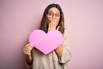 Young beautiful brunette romantic woman holding big heart paper celebrating valentine day cover mouth with hand shocked with shame for mistake, expression of fear, scared in silence, secret concept