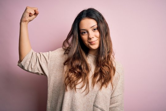 Young beautiful brunette woman wearing casual sweater standing over pink background Strong person showing arm muscle, confident and proud of power