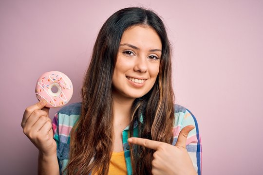 Young beautiful brunette woman eating sweet pink doughnut over isolated background with surprise face pointing finger to himself