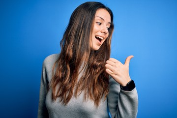 Fototapeta premium Young beautiful brunette woman wearing casual sweater standing over blue background smiling with happy face looking and pointing to the side with thumb up.
