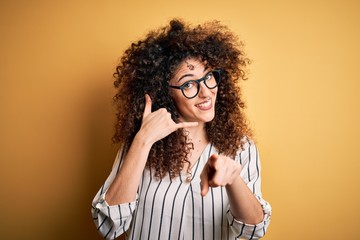 Young beautiful woman with curly hair and piercing wearing striped shirt and glasses smiling doing...