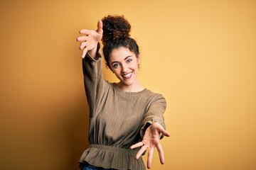 Young beautiful brunette woman with curly hair and piercing wearing casual dress looking at the camera smiling with open arms for hug. Cheerful expression embracing happiness. - obrazy, fototapety, plakaty