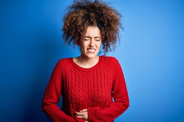 Fototapeta na wymiar Young beautiful woman with curly hair and piercing wearing casual red sweater with hand on stomach because nausea, painful disease feeling unwell. Ache concept.