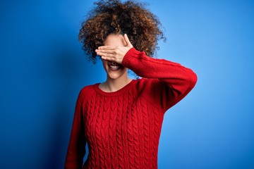Fototapeta na wymiar Young beautiful woman with curly hair and piercing wearing casual red sweater smiling and laughing with hand on face covering eyes for surprise. Blind concept.