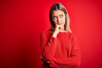 Young beautiful blonde woman wearing casual sweater over red isolated background with hand on chin thinking about question, pensive expression. Smiling with thoughtful face. Doubt concept.