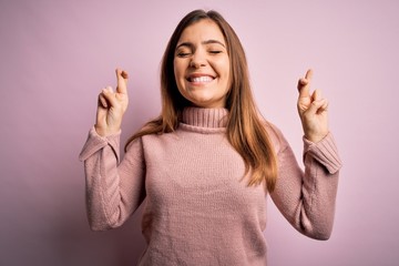 Beautiful young woman wearing turtleneck sweater over pink isolated background gesturing finger crossed smiling with hope and eyes closed. Luck and superstitious concept.