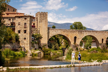 Fototapeta na wymiar Young mother with her teenager son walking near old bridge over the river Fluvia in unique medieval town of Besalu, province Girona, Spain
