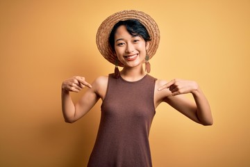 Young beautiful asian girl wearing casual t-shirt and hat over isolated yellow background looking...