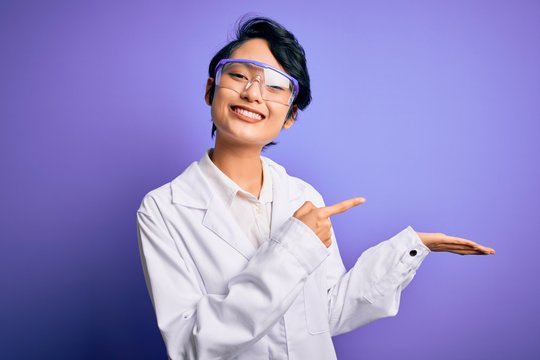 Young beautiful asian scientist girl wearing coat and glasses over purple background amazed and smiling to the camera while presenting with hand and pointing with finger.