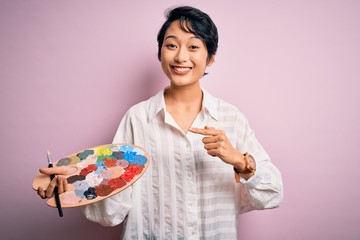 Young beautiful chinese artist woman painting using paintbrush and palette with colors very happy...