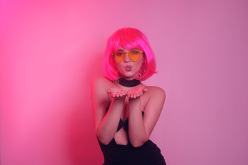 Bright girl on a pink background, in a pink short wig with bangs. The girl sends a kiss to the camera. Bright summer photo of a girl. Sweet photo of model on pink background. Joy and happiness.