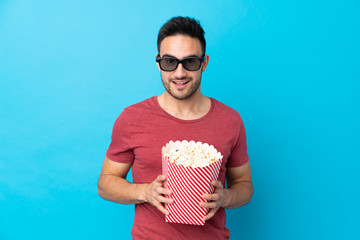 Young handsome man over isolated blue background with 3d glasses and holding a big bucket of...