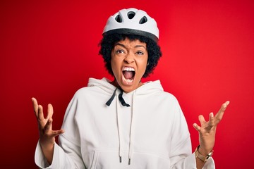 Young African American afro cyciling woman with curly hair wearing bike security helmet crazy and mad shouting and yelling with aggressive expression and arms raised. Frustration concept.