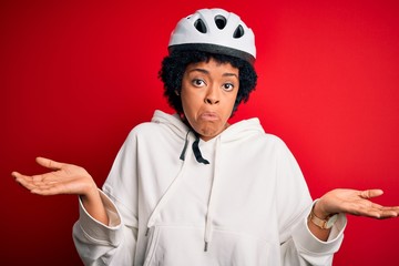 Young African American afro cyciling woman with curly hair wearing bike security helmet clueless and confused expression with arms and hands raised. Doubt concept.