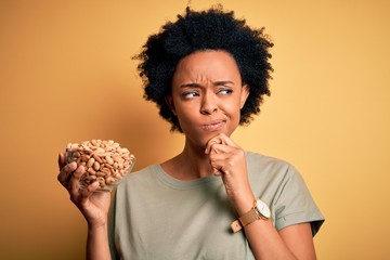 Young African American afro woman with curly hair holding bowl with salty peanuts serious face...