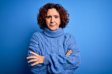 Middle age beautiful curly hair woman wearing casual turtleneck sweater over blue background skeptic and nervous, disapproving expression on face with crossed arms. Negative person.