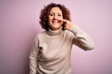 Middle age beautiful curly hair woman wearing casual turtleneck sweater over pink background Pointing with hand finger to face and nose, smiling cheerful. Beauty concept