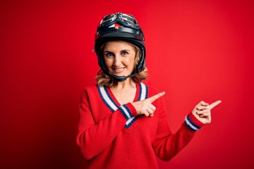 Middle age beautiful blonde motorcyclist woman wearing moto helmet over red background smiling and looking at the camera pointing with two hands and fingers to the side.