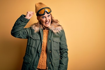 Middle age beautiful blonde skier woman wearing snow sportwear and ski goggles angry and mad raising fist frustrated and furious while shouting with anger. Rage and aggressive concept.
