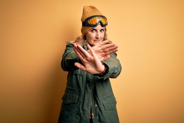 Middle age beautiful blonde skier woman wearing snow sportwear and ski goggles Rejection expression crossing arms and palms doing negative sign, angry face